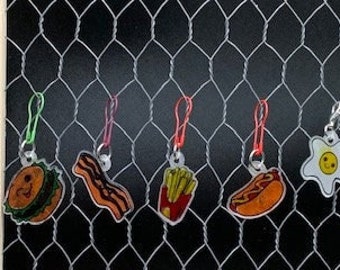 May include: Set of six colorful zipper pulls featuring various food items, including a burger, bacon, french fries, a hot dog, and a fried egg. Each zipper pull has a smiling face.