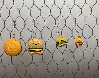 May include: Four different sized hamburger charm keychains hanging from a wire mesh background. The charms are made of plastic and feature a yellow bun, a brown patty, lettuce, and a tomato. The smallest charm has a simple design, while the largest charm has a smiling face.