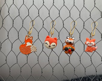 May include: Four fox-themed stitch markers with gold metal loops. The markers are made of polymer clay and feature different fox designs, including a curled-up fox, a fox with a berry, and a fox with a closed eye.