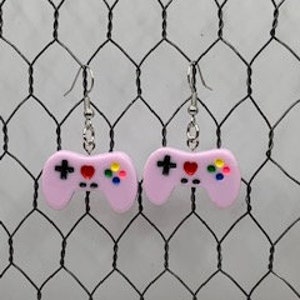 May include: A pair of pink video game controller earrings with silver-colored hooks. The controllers have black directional pads, red hearts, and colorful buttons. The earrings are displayed against a white wire mesh background.