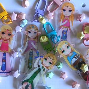 以下が含まれることがあります： 金髪のキャラクターがピンク、青、緑の衣装を着たアクリルキーホルダーのコレクション。各キーホルダーにはカラフルな留め金が付いており、折り紙の星に囲まれています。