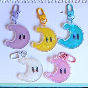 Op de afbeelding: Vijf maanvormige sleutelhangers in paars, geel, blauwgroen, wit en roze. Elke sleutelhanger heeft een cartoongezicht met twee zwarte ovale ogen. De sleutelhangers hebben metalen clips om aan tassen of sleutels te bevestigen.