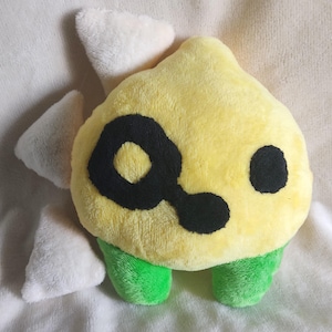 Puede incluir: Un juguete de peluche amarillo con ojos negros y un diseño de espiral negro. El juguete tiene pies verdes y orejas blancas puntiagudas.