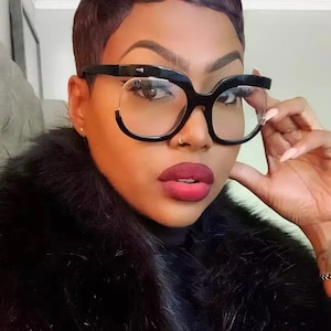 May include: Oversized black spectacles featuring a thick, bold frame design with a distinct cut-out detail along the lower rim. The glasses have clear lenses and a glossy finish, providing a prominent look that frames the face with a solid black structure.