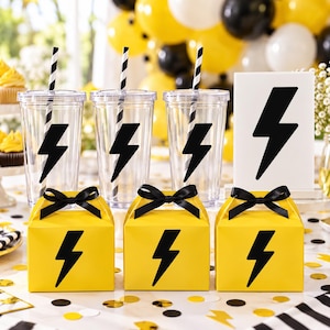 Könnte beinhalten: Drei durchsichtige Kunststoffbecher mit schwarz-weiß gestreiften Strohhalmen, jeweils mit einem schwarzen Blitz-Aufkleber. Drei gelbe Geschenkboxen mit schwarzen Schleifen und schwarzen Blitz-Symbolen sowie ein weißes Schild mit einem schwarzen Blitz.