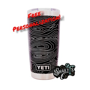 May include: A black Yeti tumbler with a white topographic pattern design. The tumbler has a silver lid and the word "YETI" is printed on the side. The text "FREE PERSONALIZATION!" is written in red on the top of the tumbler.