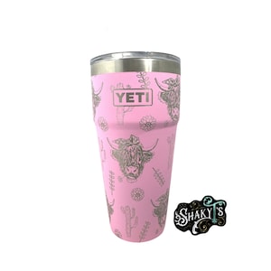 May include: Pink Yeti tumbler with a silver lid and a design of Highland cows and cacti. The tumbler has the word "YETI" printed on the side.
