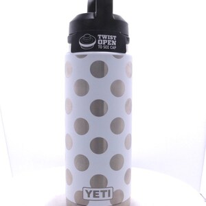 May include: A white Yeti Rambler with a silver polka dot pattern and a black lid. The lid has a black handle and a sticker that says "Twist Open to See Cap".