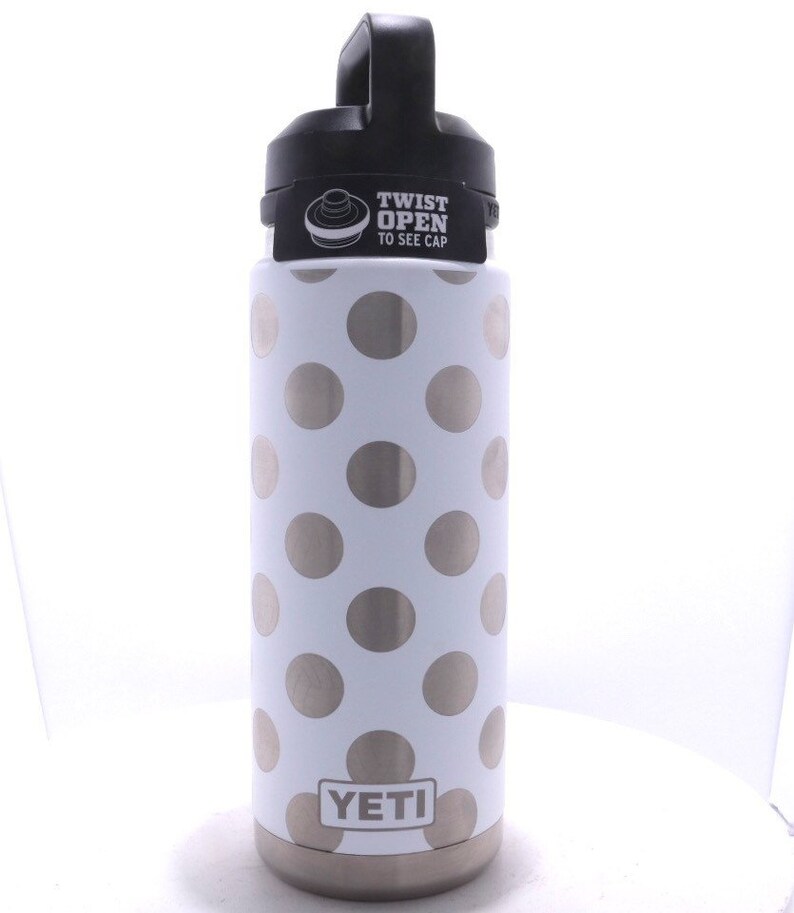 May include: A white Yeti Rambler with a silver polka dot pattern and a black lid. The lid has a black handle and a sticker that says "Twist Open to See Cap".