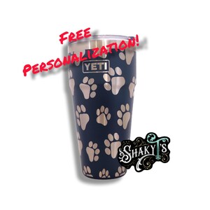 May include: A navy blue Yeti tumbler with a paw print design. The tumbler has a silver lid and the Yeti logo is visible. The text "FREE PERSONALIZATION!" is written in red above the tumbler.