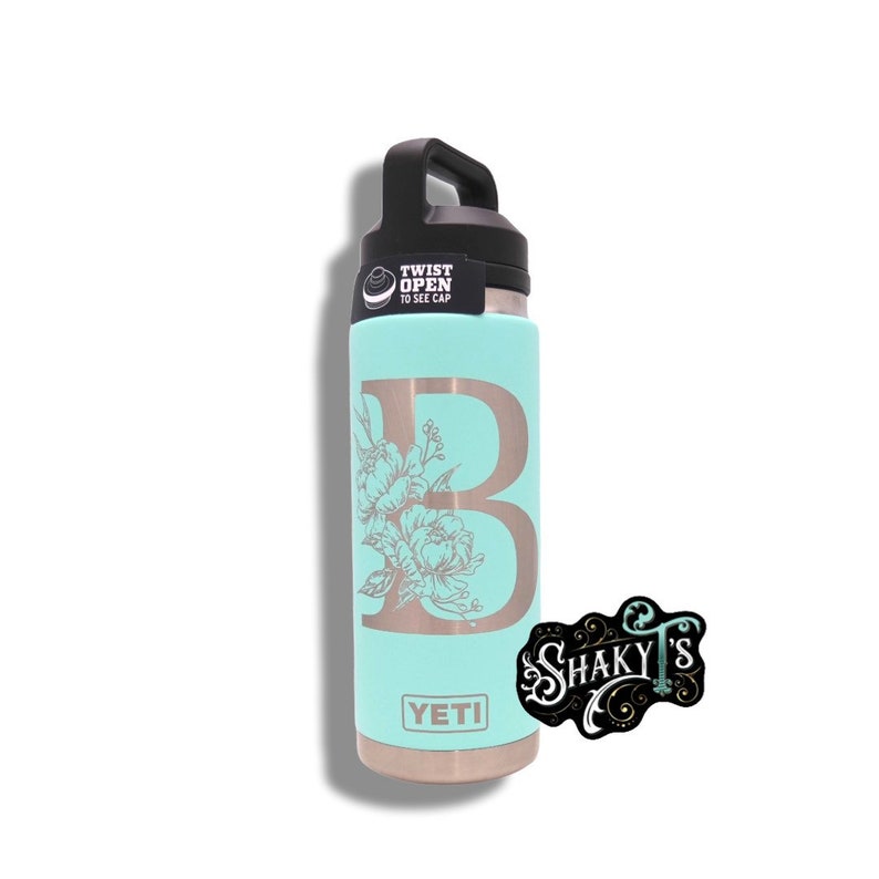 May include: A turquoise Yeti water bottle with a black lid and a silver twist-open cap. The bottle has a floral design and the letter "B" engraved on the side. The bottle has the word "YETI" printed on the bottom.