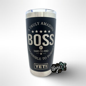 May include: A black Yeti tumbler with a silver rim and lid. The tumbler features the text "A Truly Amazing BOSS is Hard to Find, Impossible to Forget" in silver lettering. The Yeti logo is at the bottom. A small "SHAKY'S" logo is in the bottom right corner.