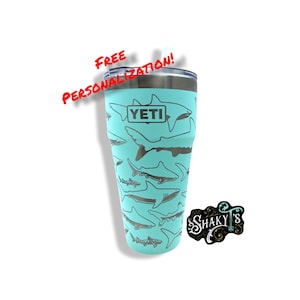 Peut inclure: Un YETI Rambler turquoise avec un motif de requins. Le gobelet a un couvercle et une paille. Le texte "FREE PERSONALIZATION!" est écrit en rouge au-dessus du gobelet. Un autocollant noir avec le texte "SHAKY'S" est dans le coin inférieur droit de l'image.