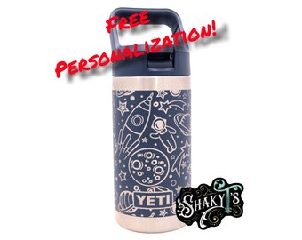 May include: A stainless steel water bottle with a navy blue lid and a space-themed design. The bottle has a black and white design of stars, planets, rockets, and astronauts. The bottle has the word "YETI" printed on it. The bottle is sitting on a white background. "FREE PERSONALIZATION!" is written in red text above the bottle.