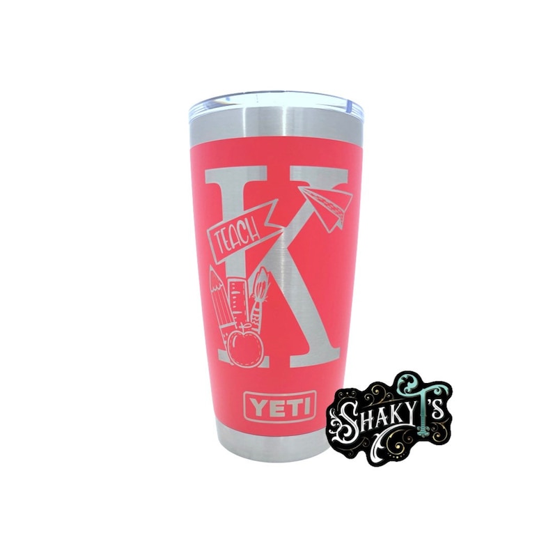 May include: A pink YETI tumbler with a silver lid and a silver K with the word "TEACH" on a banner above it. There are school supplies like a pencil, ruler, paintbrush, and apple around the K. The words "SHAKY'S" are on a black sticker with a white design.