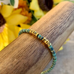 May include: A turquoise and gold beaded bracelet with a delicate design. The bracelet is made of small, round beads with a subtle shimmer. The gold beads are accented with a geometric pattern.