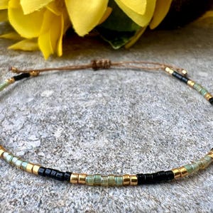 Könnte beinhalten: Ein zartes Armband mit schwarzen, goldenen und grünen Perlen. Das Armband hat einen einfachen Verschluss aus Schnur.