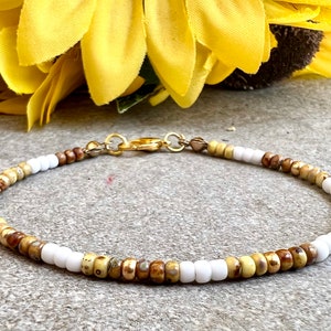 Op de afbeelding: Een delicate armband gemaakt met kleine witte, bruine en gouden kralen. De armband heeft een gouden sluiting.
