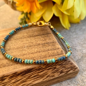 Pode incluir: Delicada pulseira de contas com uma mistura de contas azuis e turquesas, intercaladas com pequenas contas douradas. A pulseira tem um fecho dourado e &eacute; exibida sobre uma superf&iacute;cie de madeira, com flores amarelas ao fundo.