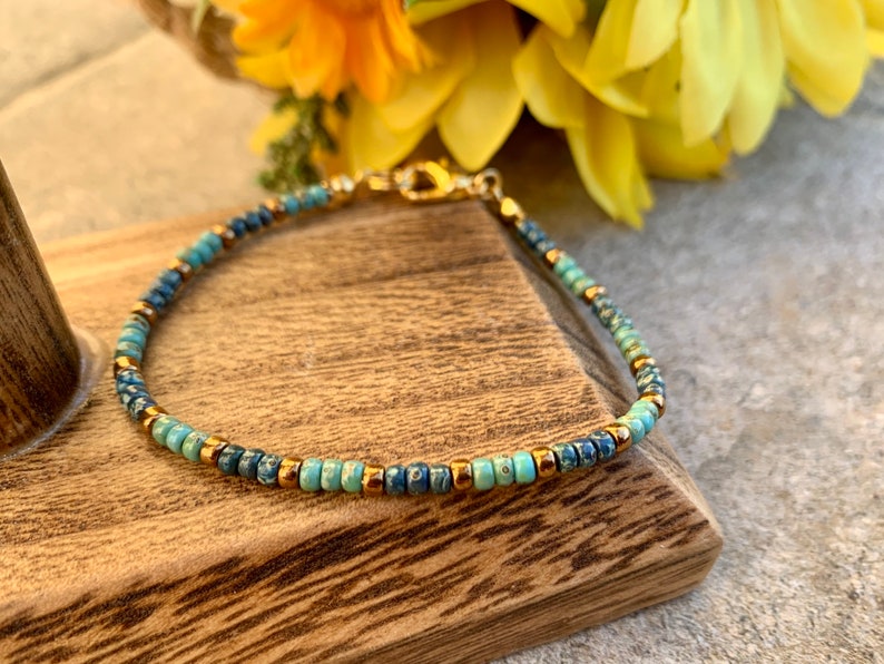 Pode incluir: Delicada pulseira de contas com uma mistura de contas azuis e turquesas, intercaladas com pequenas contas douradas. A pulseira tem um fecho dourado e &eacute; exibida sobre uma superf&iacute;cie de madeira, com flores amarelas ao fundo.