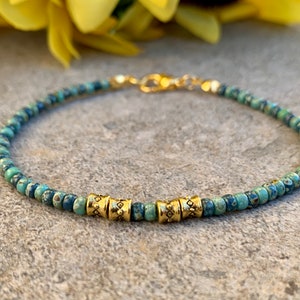 May include: A delicate bracelet made with small blue and green patterned beads and gold accents. The bracelet has a simple design and is perfect for everyday wear.