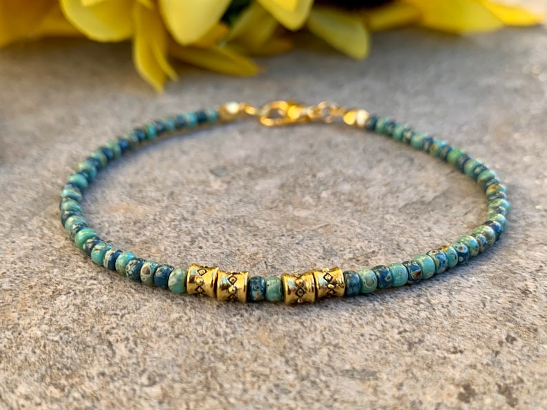 May include: A delicate bracelet made with small blue and green patterned beads and gold accents. The bracelet has a simple design and is perfect for everyday wear.