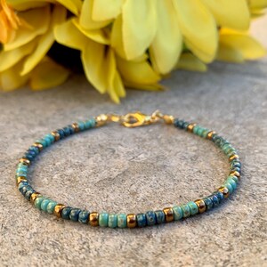 May include: Delicate beaded bracelet featuring a mix of blue and turquoise beads with gold-colored accents. The bracelet is strung on a gold-colored chain and has a clasp closure. A stylish piece of jewelry.