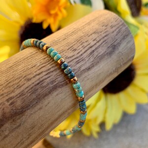 Pode incluir: Uma pulseira de contas com contas turquesa e azul escuro, intercaladas com pequenas contas douradas. A pulseira &eacute; exibida em um pino de madeira, com girass&oacute;is ao fundo. Uma delicada pe&ccedil;a de joalheria.