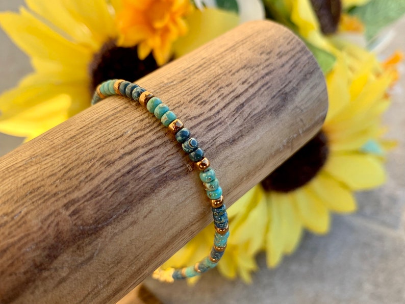 Pode incluir: Uma pulseira de contas com contas turquesa e azul escuro, intercaladas com pequenas contas douradas. A pulseira &eacute; exibida em um pino de madeira, com girass&oacute;is ao fundo. Uma delicada pe&ccedil;a de joalheria.