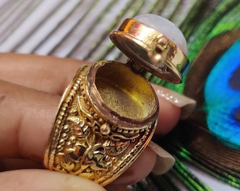 Può includere: Un anello d'oro con un designo ornato e un coperchio incernierato. Il coperchio è aperto e rivela un piccolo scomparto vuoto. L'anello è incastonato con una pietra bianca.