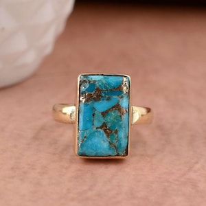 May include: A gold ring with a rectangular turquoise stone set in the center. The stone has a blue and green color with brown flecks throughout.