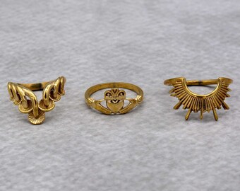 Può includere: Tre anelli dorati. L'anello a sinistra ha un design delicato e traforato. L'anello centrale presenta un motivo a nodo celtico con un cuore al centro. L'anello a destra ha un design a raggiera.