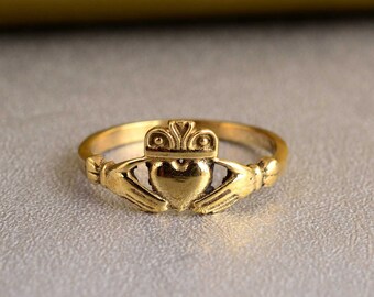 Può includere: Un anello Claddagh dorato con un design a forma di cuore, due mani che stringono il cuore e una corona in cima.