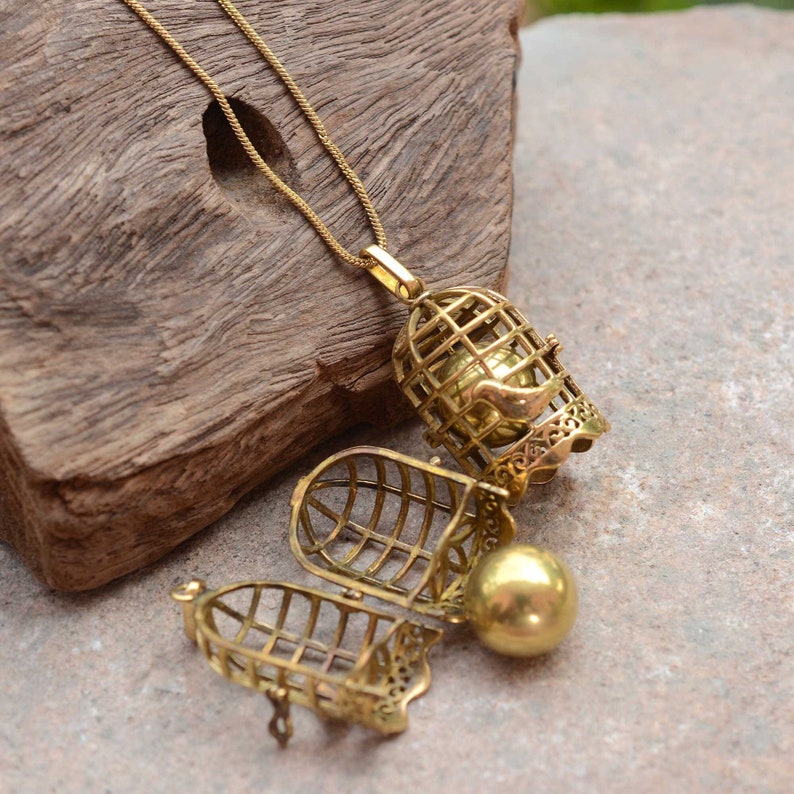 Puede incluir: Un collar con un colgante de jaula de tono dorado con una peque&ntilde;a bola de oro en su interior. La jaula est&aacute; abierta y la bola es visible. El collar est&aacute; sobre una superficie de madera.