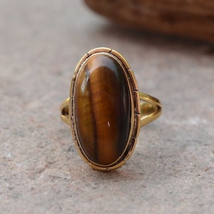 Könnte beinhalten: Ein goldener Ring mit einem ovalen Tigerauge-Edelstein. Der Ring hat ein schlichtes Band und eine Zargenfassung.