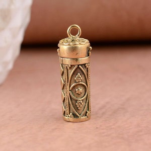 Puede incluir: Un colgante de metal dorado en forma de un pequeño cilindro adornado con una tapa con bisagras. El cilindro está decorado con intrincados patrones geométricos y motivos florales.