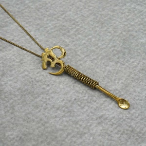 May include: A gold necklace with a pendant shaped like the Om symbol. The pendant is attached to a long, thin, gold chain. The pendant has a small spoon at the end.