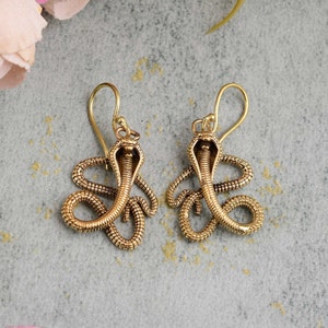 Peut inclure: Une paire de boucles d'oreilles en forme de serpent dorées. Les boucles d'oreilles sont en métal et présentent un motif de serpent détaillé avec une tête surélevée et un corps enroulé.