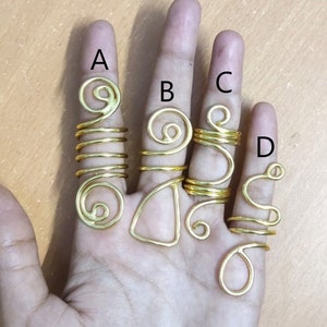 Könnte beinhalten: Vier goldfarbene Haarreifen aus Metall, mit den Buchstaben A, B, C und D gekennzeichnet, sind auf einer Hand zu sehen. Die Ringe haben unterschiedliche Formen und Größen, einige haben ein Spiraldesign.