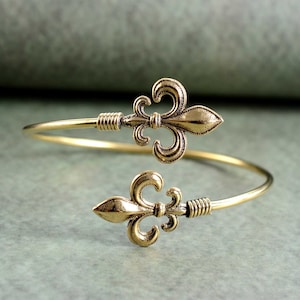 May include: A gold-toned metal bracelet with two fleur-de-lis charms. The bracelet is open and adjustable.