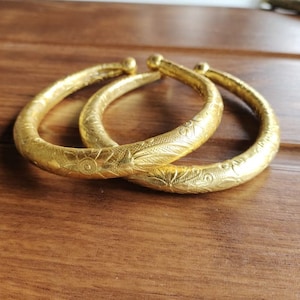 May include: Two gold-coloured bangles with intricate floral designs. The bangles are stacked on top of each other.