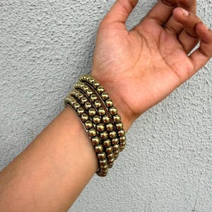 May include: A stack of four gold-toned beaded bracelets featuring uniform, polished spherical beads. The bracelets are worn together, creating a layered appearance with a metallic, reflective finish on each individual bead in the set.