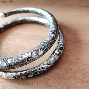 May include: Two silver bangles with intricate floral designs. The bangles are stacked on top of each other and have a textured surface.
