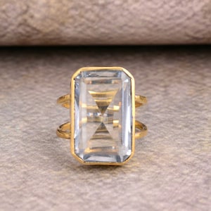 May include: A gold ring featuring a large, clear, rectangular gemstone. The gemstone is faceted and has a subtle blue hue.