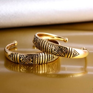 May include: Two gold-toned bangles with intricate geometric patterns. The bangles are stacked on top of each other.