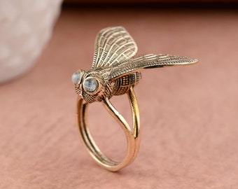 Può includere: Un anello color oro con un designo di ape dettagliato con occhi di pietre preziose trasparenti. Le ali dell'ape sono spiegate, conferendo all'anello un aspetto unico e fantasioso.