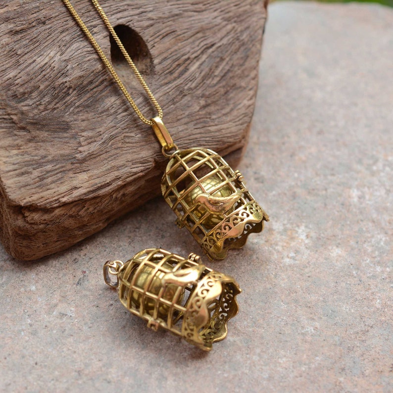 Puede incluir: Dos colgantes de jaula de p&aacute;jaros de tono dorado con un peque&ntilde;o p&aacute;jaro dentro. El colgante m&aacute;s grande est&aacute; en una cadena dorada.