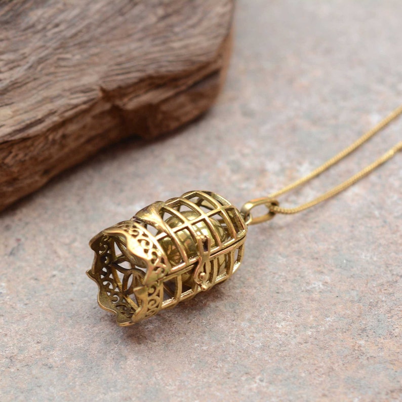 Puede incluir: Un collar dorado con un colgante en forma de jaula que contiene una peque&ntilde;a bola. El colgante est&aacute; intrincadamente detallado y tiene un dise&ntilde;o delicado.