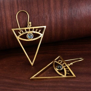 May include: A pair of gold triangle earrings with a blue eye design. Each earring features a triangle shape with a detailed eye design and a blue gemstone in the center.