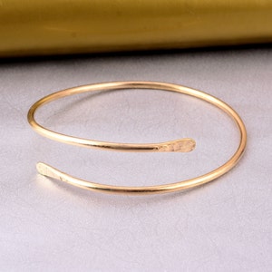May include: A gold-toned, open-ended bangle bracelet with a simple, minimalist design. The bracelet is made of a single, continuous piece of metal that is bent into a circle.